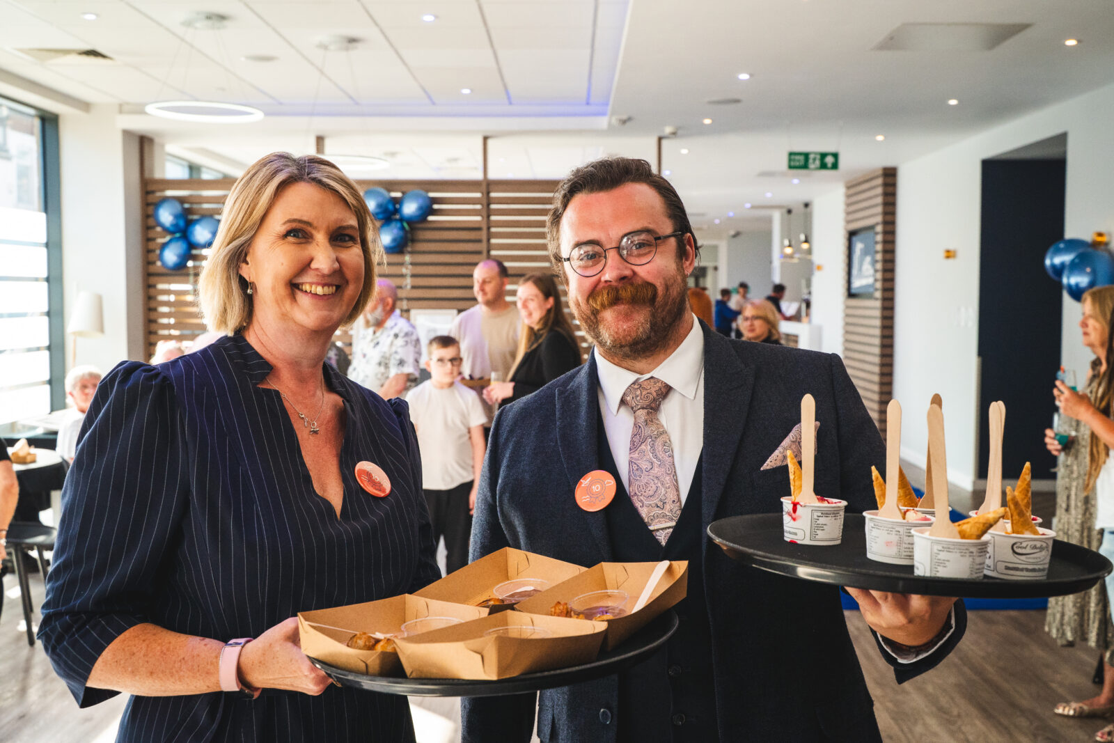 A man and a woman in suits smiling and holding food on a tray